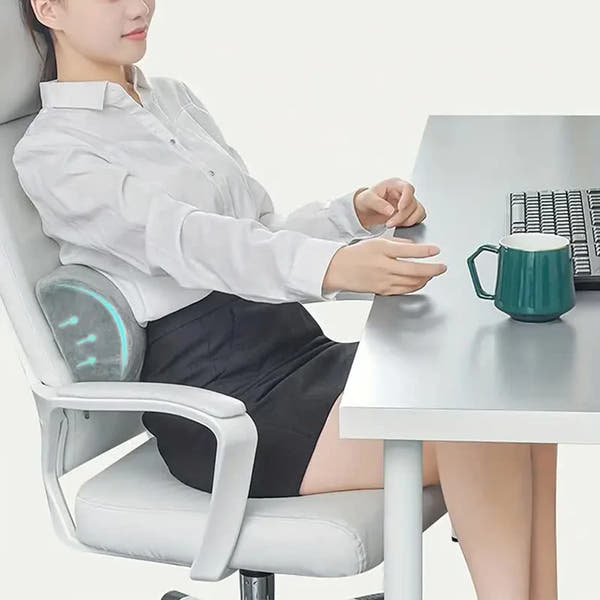 Femme assise au bureau utilisant un coussin lombaire gris demi-lune placé entre le dossier et le bas du dos.